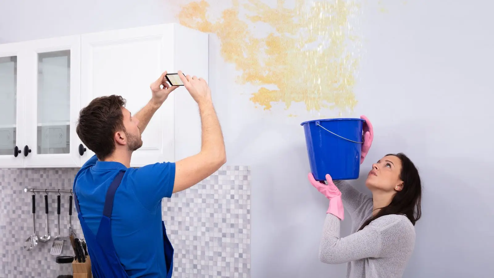 Couple photographing a leaking kitchen ceiling while catching drips in a bucket