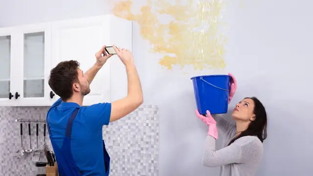 Couple photographing a leaking kitchen ceiling while catching drips in a bucket
